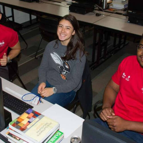 Three students are seated at a computer desk, smiling at the camera in a classroom or lab setting. Two of them, wearing matching red shirts with white logos, sit on either side of a woman in a gray sweatshirt with a hornet graphic; books and a computer.